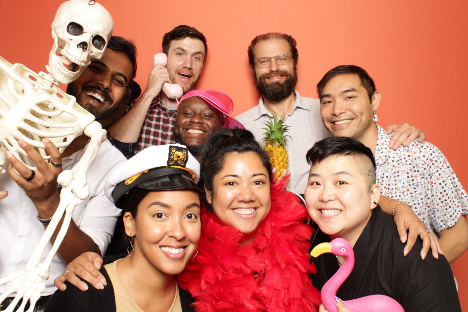 Haven Technologies employees posing for a photo with a number of silly props. On the far left, a man is holding a plastic skeleton. In the top center, one is on a corded phone, with the man on his right holding a pineapple. On the bottom left, a woman is wearing a captain's hat. The woman next to her is in a pink feather boa, and the person on that woman's right is holding a prop flamingo.