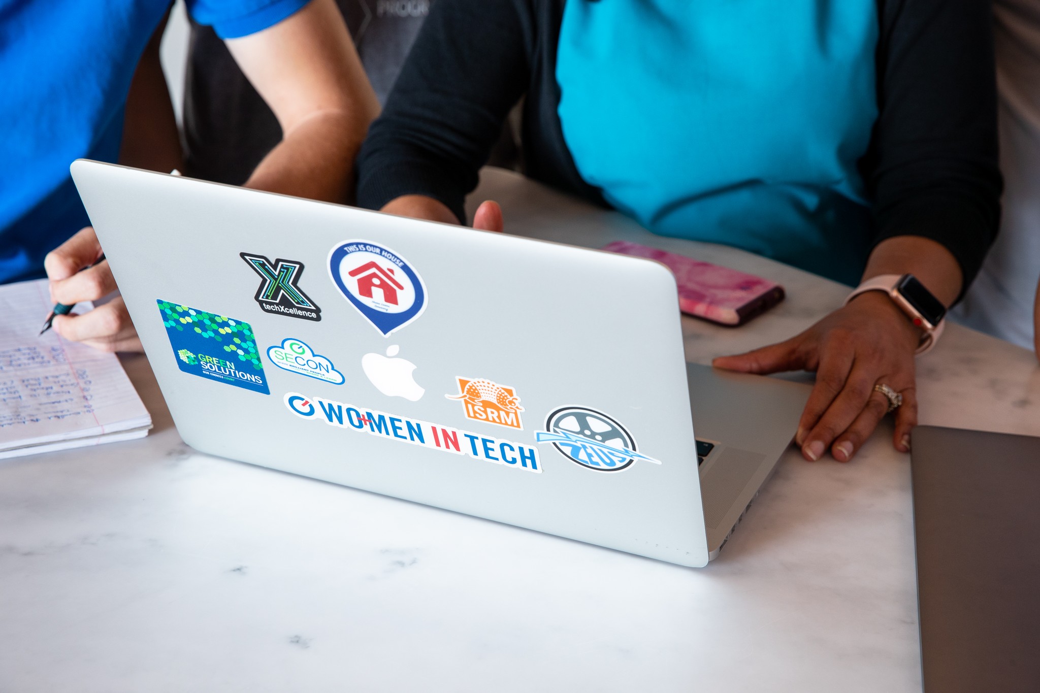 Close-up photo of the back of a laptop with stickers on it and two people seated next to each other facing the screen.