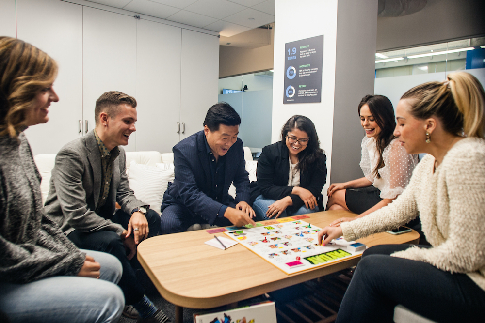 Team members at DailyPay gather around a table while laughing and playing a board game together.