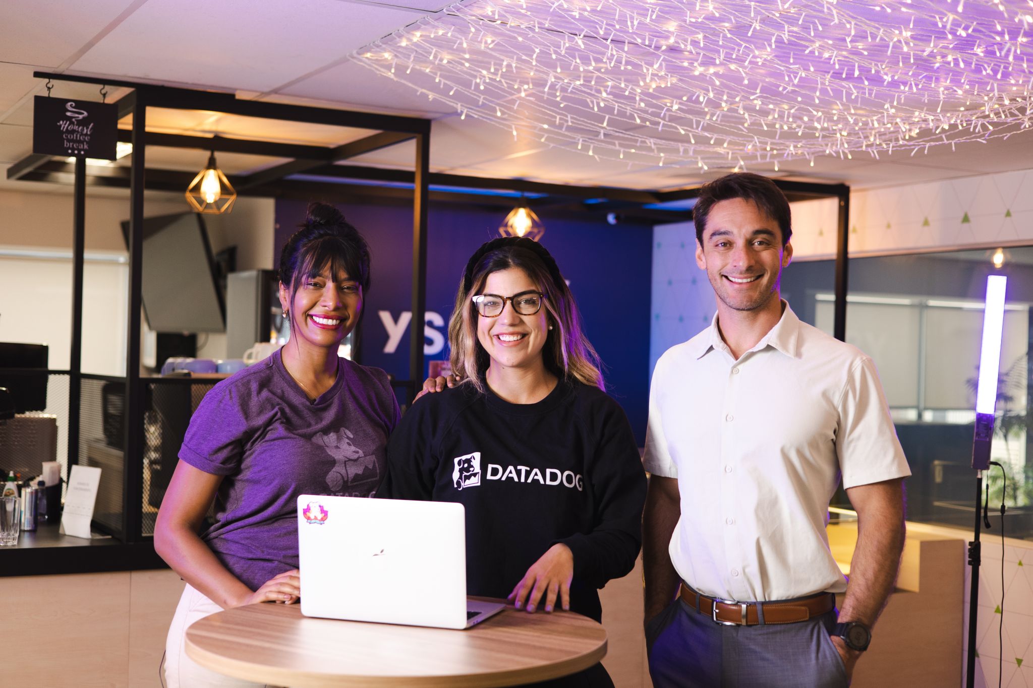 Three Datadog team members smile for a group photo in the office, a laptop in front of them. 