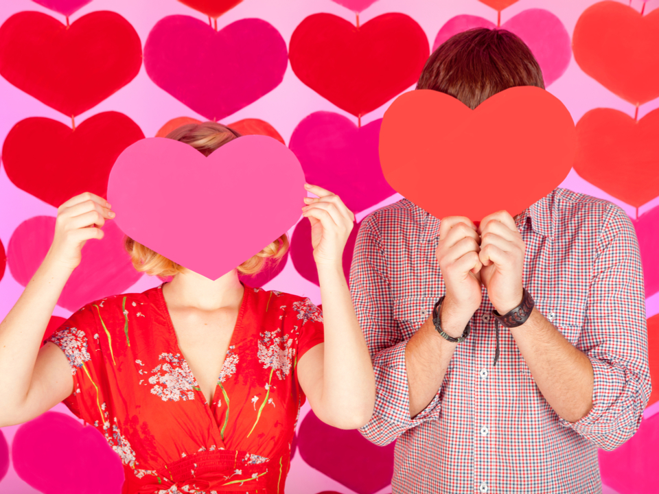 Two people hold red paper hearts over their faces against a romantic background