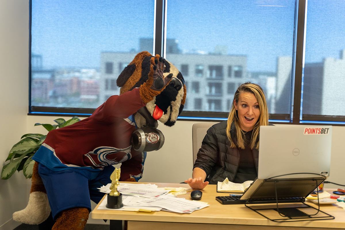 PointsBet team member sitting at a desk working on a laptop with the person in a dog costume wearing an Avalanche jersey in her office