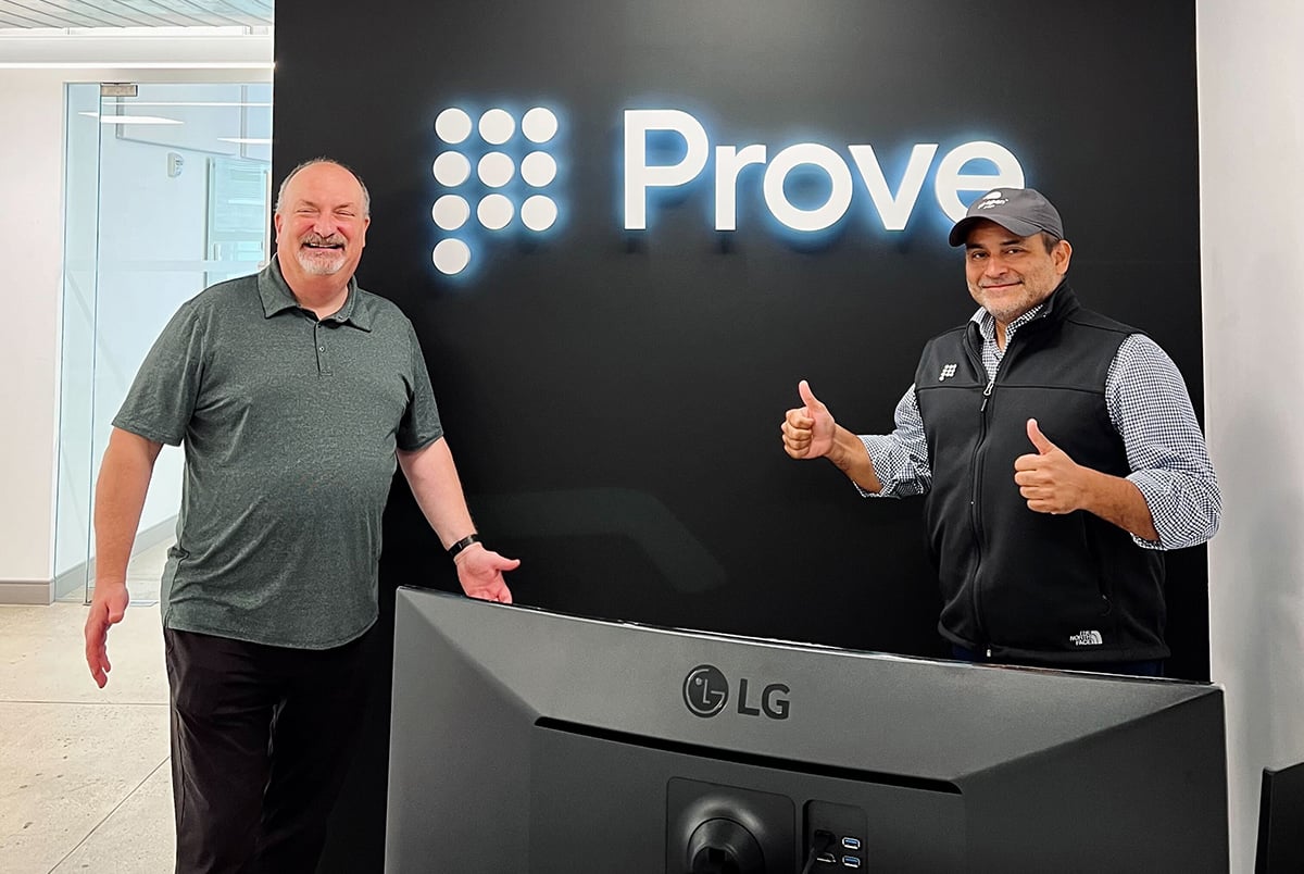 Two Prove team members standing in front of the Prove logo on a wall in the office