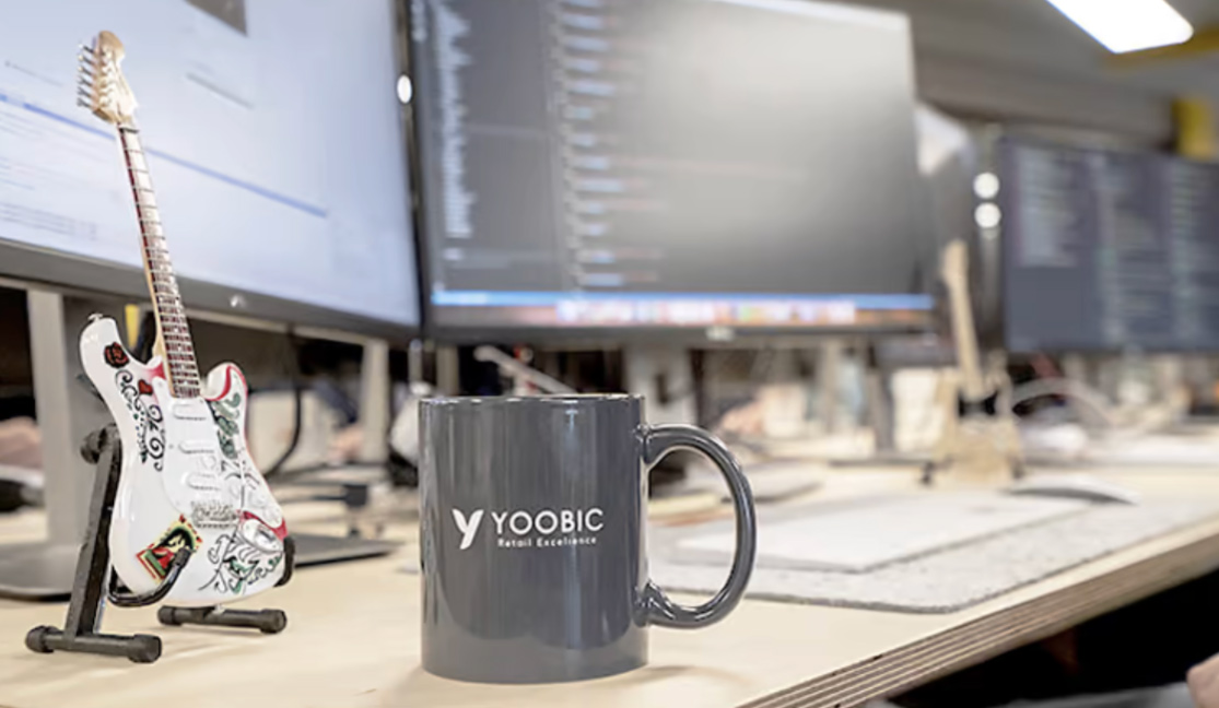 Mug with the YOOBIC logo on it on a desk with a mini guitar next to it and computer monitors in the backgroud