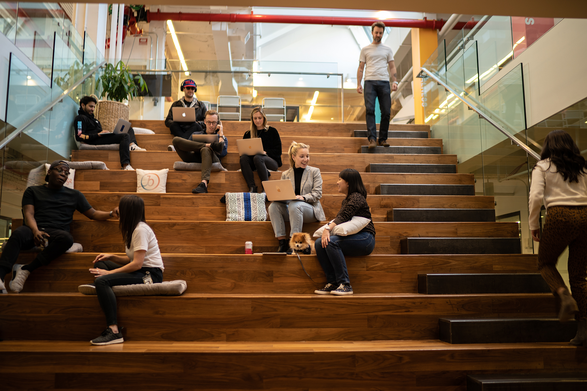 Employees sit with laptops along large wooden staircase, talking while working.