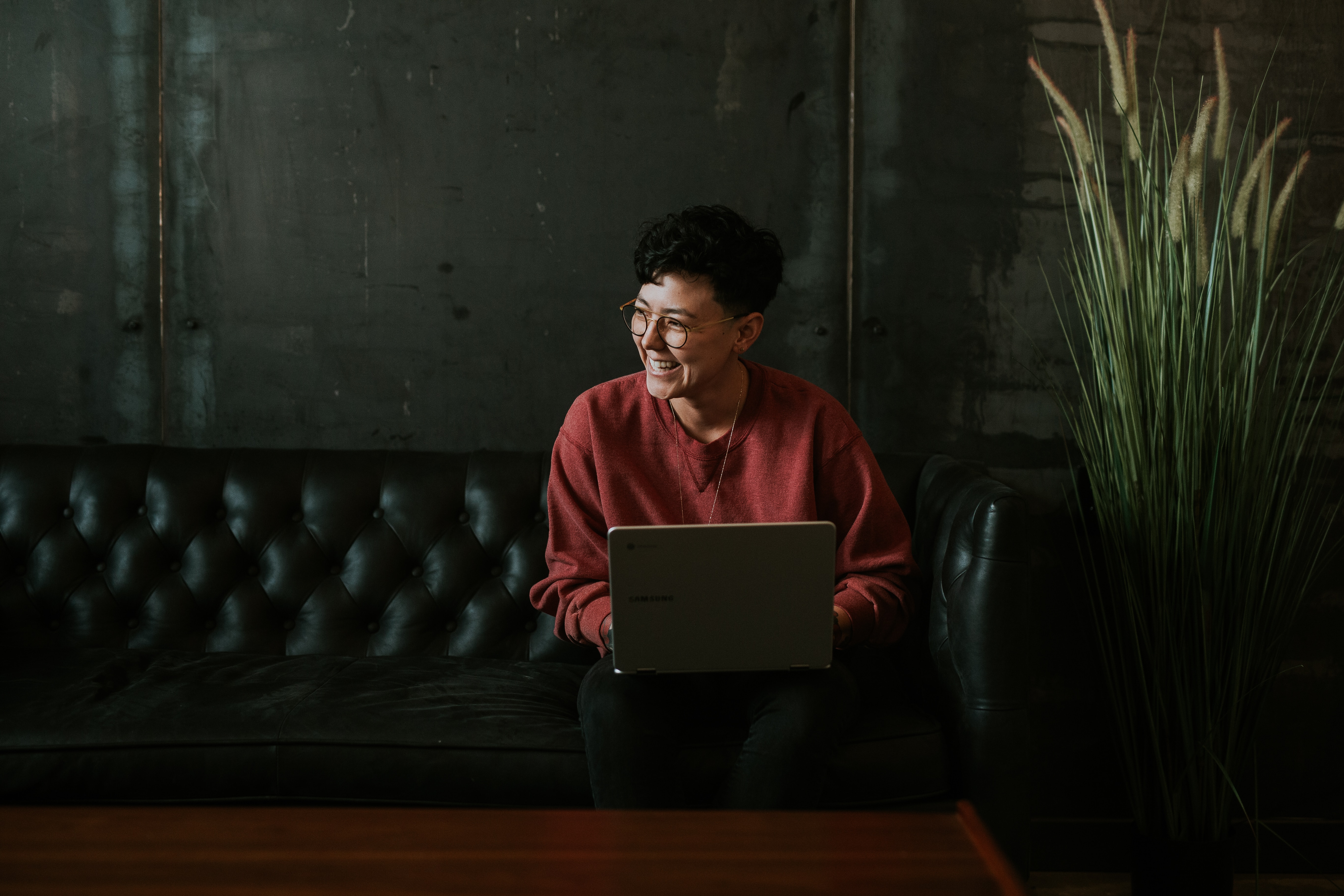 Unsplash photo: Dark room with woman on leather couch next to a tall plant, laptop on lap, laughing