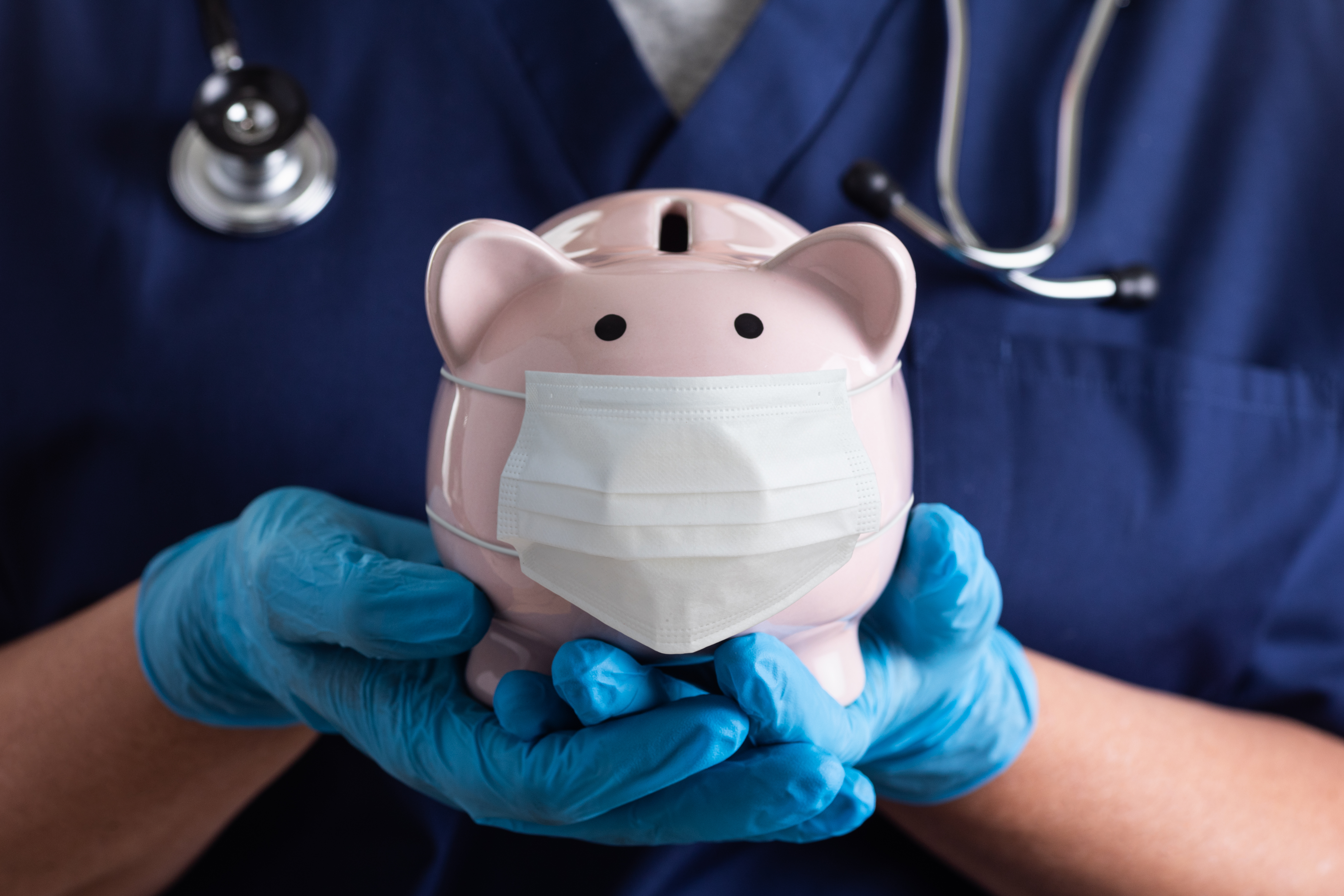 A doctor wearing surgical gloves holds a piggy bank wearing a face mask. 