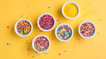 Image of six ceramic jars containing various types of colorful sprinkles on a yellow background, from a bird's eye view