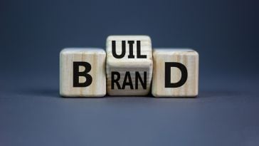 Three building blocks in a line. The first is a B, the last is a D. In the middle, it is tilted between "uil" (making "build") and "ran" (making "brand)"