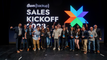 Photo of OwnBackup team members on a stage with large screen behind reading “OwnBackup Sales Kickoff 2023.