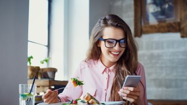 A woman eating a salad and looking at her phone is pictured.