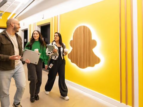 Snap team members smiling and talking while walking through office hallway featuring yellow wall with illuminated Snap logo